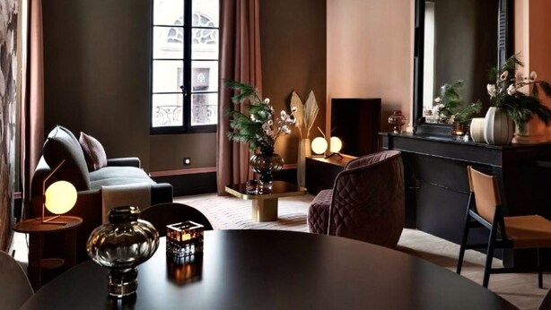 A modern hotel room with a dark brown wall, a large window with curtains, a round table with a lamp, a sofa, and a mirror. - Notre-Dame 3* - OG IMMO (Annecy)