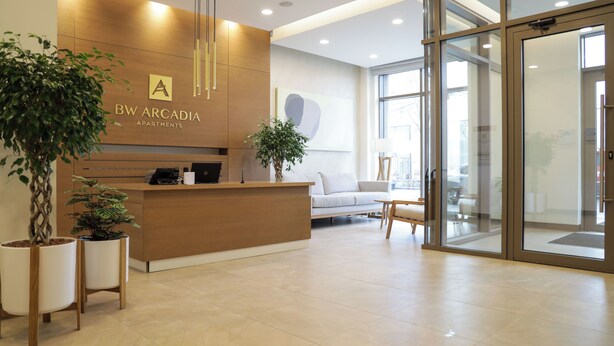 A reception area with a wooden reception desk, potted plants, a sofa, and a glass door leading to another room. - Apartments Royal - Arcadia Building (Belgrade)