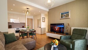 A modern living room with a sofa, a green armchair, a coffee table with fruit, a TV, and a dining area with chairs and a table. - Apartments Royal - Arcadia Building (Belgrade)