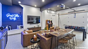 A modern living room with a foosball table, comfortable seating, a flat-screen TV, and a neon sign.