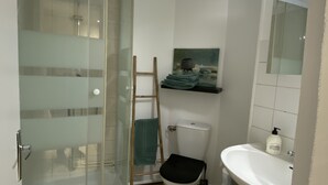A bathroom with a glass shower enclosure, a toilet, a sink, and a shelf with towels. - Studio near city center (Verdun)