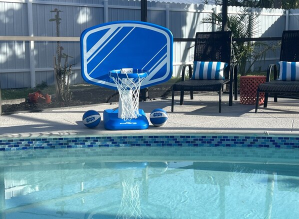 A pool area with a blue inflatable basketball hoop, lounge chairs, and a tiled pool edge.