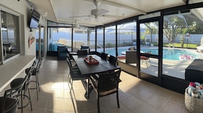 A sunlit patio with a pool view, a dining area with chairs and a table, and a ceiling fan.