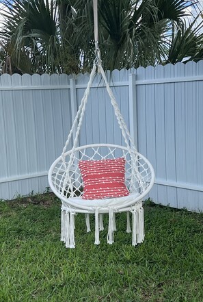 A white macrame hanging chair with a red cushion, set against a white picket fence and green grass.