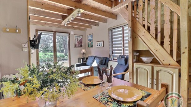 Un comedor con una mesa de madera dispuesta para dos personas, un jarrón con flores amarillas y una escalera con barandillas de madera.