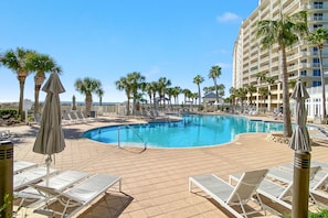 Zona de piscina con sillones de playa y sombrillas, rodeada de palmeras y un edificio de hotel alto.