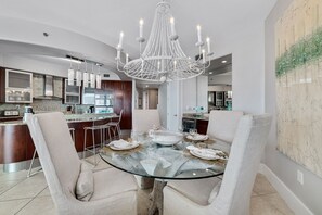 Uma área de jantar moderna com mesa de vidro, cadeiras estofadas e um lustre central.