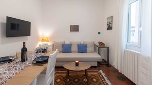 A living room with a sofa, a coffee table, a TV mounted on the wall, and a window with curtains. - Oltrarno Suite (Florence)