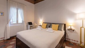 A neatly made bed with white linens and yellow pillows, a desk with a laptop, a chair, and a lamp on a bedside table. - Oltrarno Suite (Florence)
