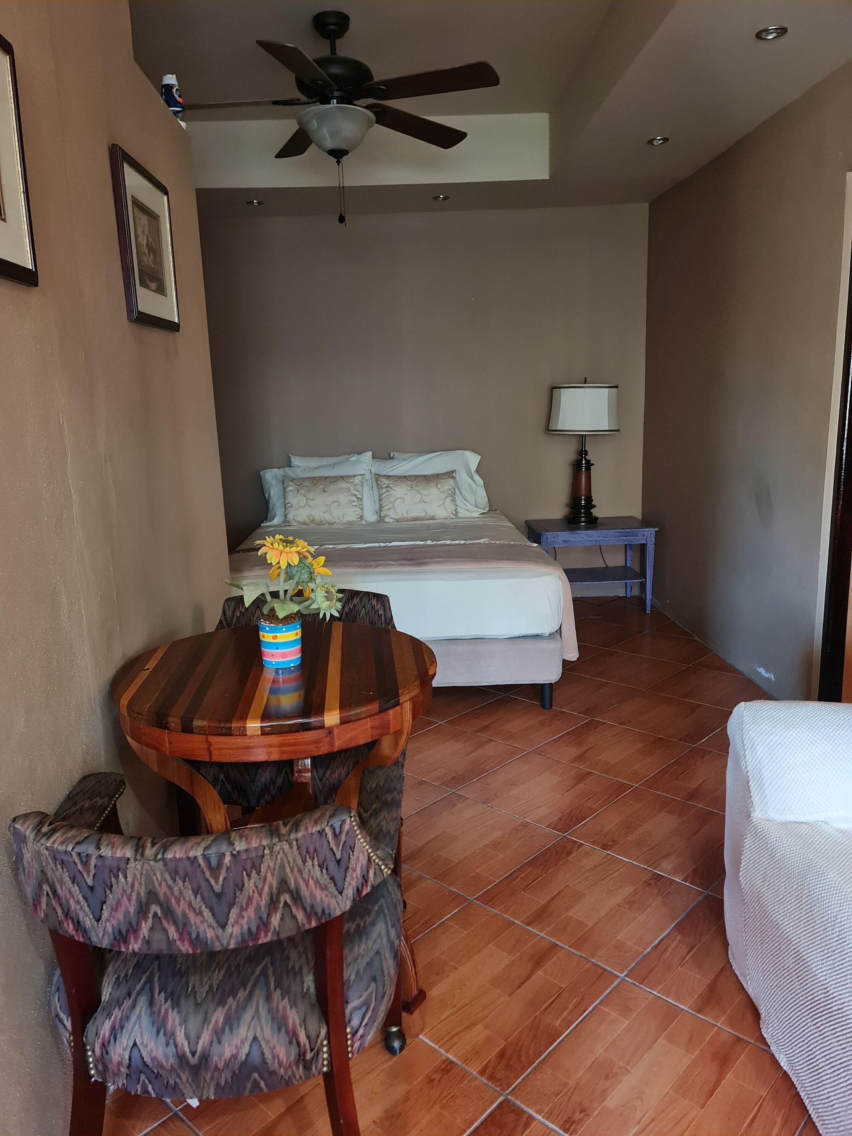 A bedroom with a bed, a ceiling fan, a bedside table with a lamp, and a small table with a vase of flowers.