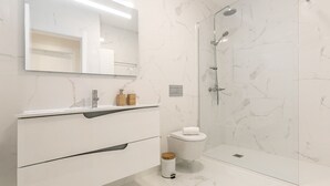 A modern bathroom with a marble floor, a glass shower enclosure, a white sink with a wooden soap dispenser, and a toilet with a white toilet paper holder.