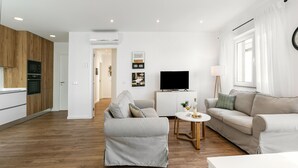 A modern living room with a sofa, a coffee table, and a television. There is a kitchen area with wooden cabinets and a microwave.