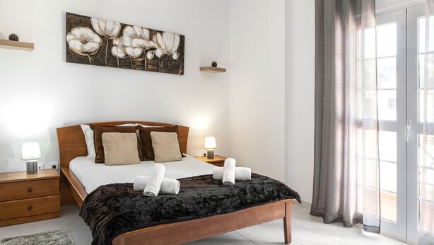 A neatly arranged bedroom with a large bed, two bedside tables, a rug, and a painting on the wall. - Relax in Foz - Shared Pool (Caldas da Rainha)