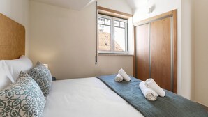 A neatly made bed with patterned pillows, a blue blanket, and two rolled towels. A window with a view of a building is visible. - Nazaré Boutique Apartment II (Nazaré)