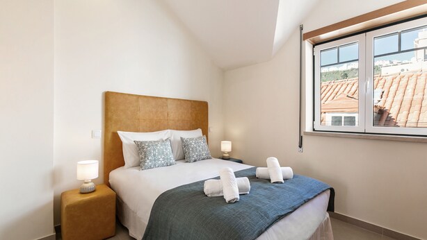 A neatly arranged hotel room with a large bed, two bedside lamps, a nightstand, and a window with a view of buildings. - Nazaré Boutique Apartment II (Nazaré)