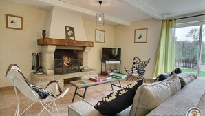A cozy living room with a stone fireplace, a television, and a sofa with patterned cushions. - Mimosa - Frehel - Gîte (FREHEL)