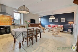 A modern kitchen with a built-in oven, a dining area with a striped tablecloth, wicker chairs, and a blue sofa in a living room with artwork on the wall. - Hippocampe - Frehel - Gîte (FREHEL)