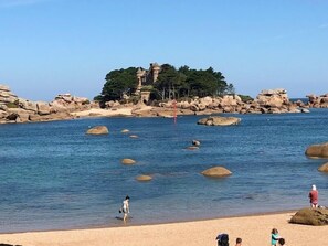 A beach with clear blue water, large rocks, and a castle-like structure in the background. - Le Linkin – PERROS GUIREC (PERROS GUIREC)