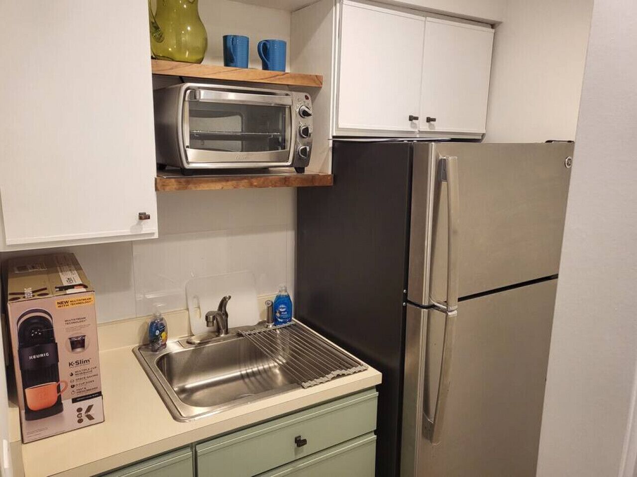 A compact kitchen with a built-in toaster oven, a stainless steel refrigerator, and a sink with a dish rack.