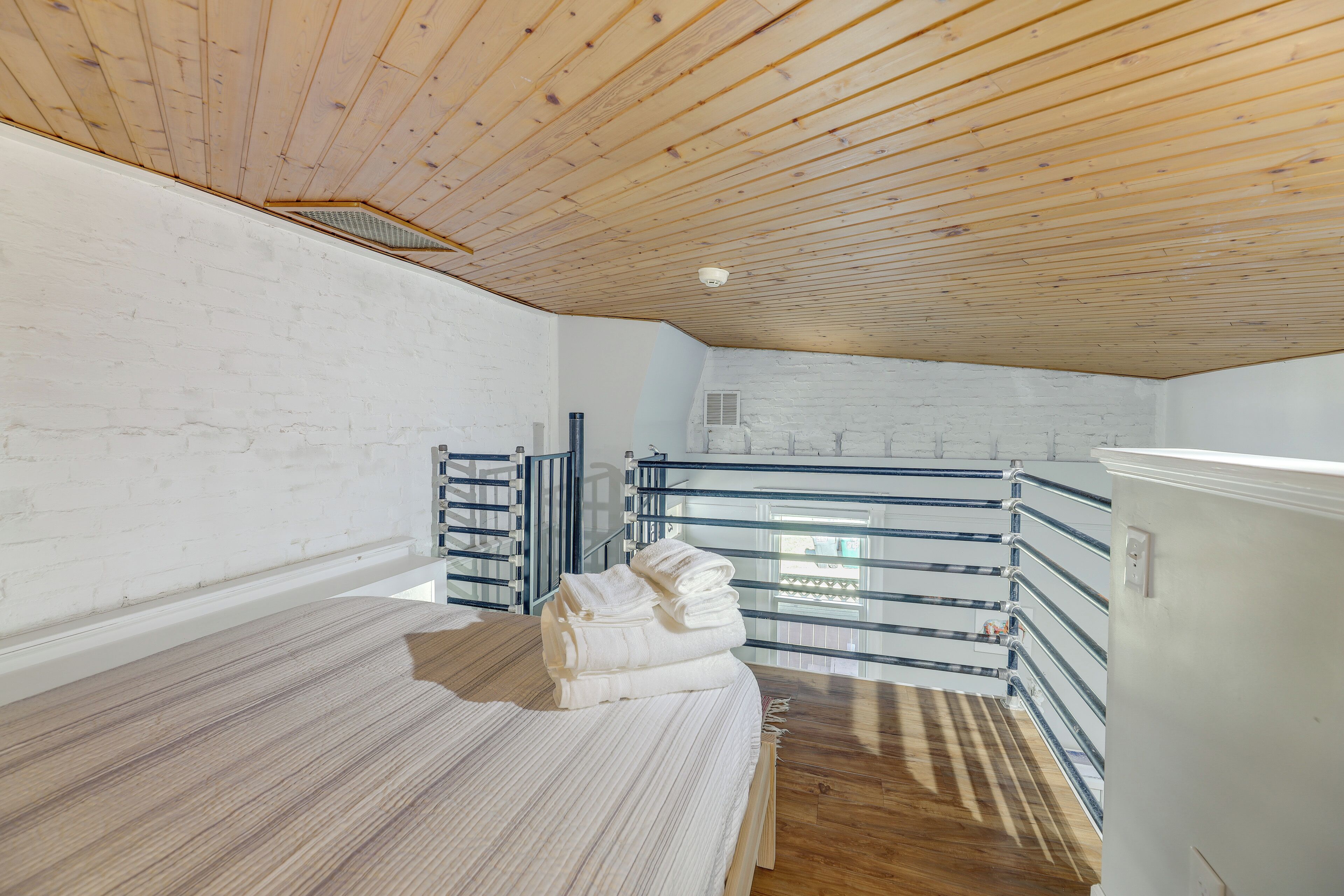 A room with a sloped ceiling, white brick walls, a bed with a striped bedspread, and a metal railing.
