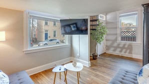 Een moderne woonkamer met een flatscreen televisie aan de muur, twee kleine witte tafels, een grijze bank en een potplant.
