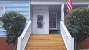 Un escalier en bois mène à une porte blanche portant le numéro 3911, de part et d’autre de laquelle se trouvent des rampes blanches et de petits arbres.