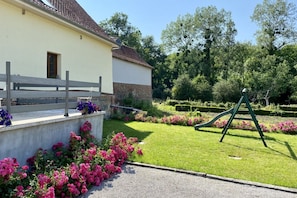 A building with a white exterior and a brick base, surrounded by a well-maintained garden with pink flowers and a green swing set. - La Grange, Ancienne Ferme Rénovée Pour 8 Personnes (Moyenneville)