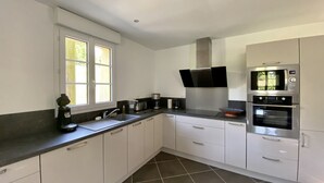 A modern kitchen with white cabinets, a stainless steel range hood, a double oven, and a window providing natural light. - La Grange, Ancienne Ferme Rénovée Pour 8 Personnes (Moyenneville)