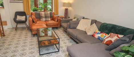 A living room with a sofa, armchairs, a coffee table, patterned floor tiles, a painting, and a window with curtains.