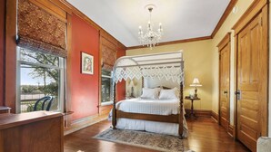 A bedroom with a four-poster bed, a chandelier, a wooden desk, and a view of the outdoors.