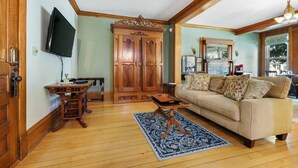 A living room with a wooden floor, a beige sofa, a wooden cabinet, and a television mounted on the wall. - Cozy king Suite, two person jet tub, Downtown! (Sturgeon Bay)
