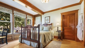 A bedroom with a wooden bed, a nightstand, a lamp, and a view of the outdoors through large windows. - Cozy king Suite, two person jet tub, Downtown! (Sturgeon Bay)
