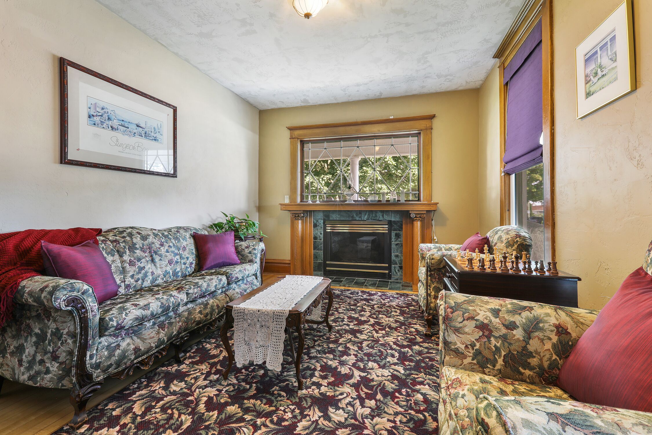A living room with a fireplace, floral-patterned sofas, a chessboard, and a framed picture on the wall.