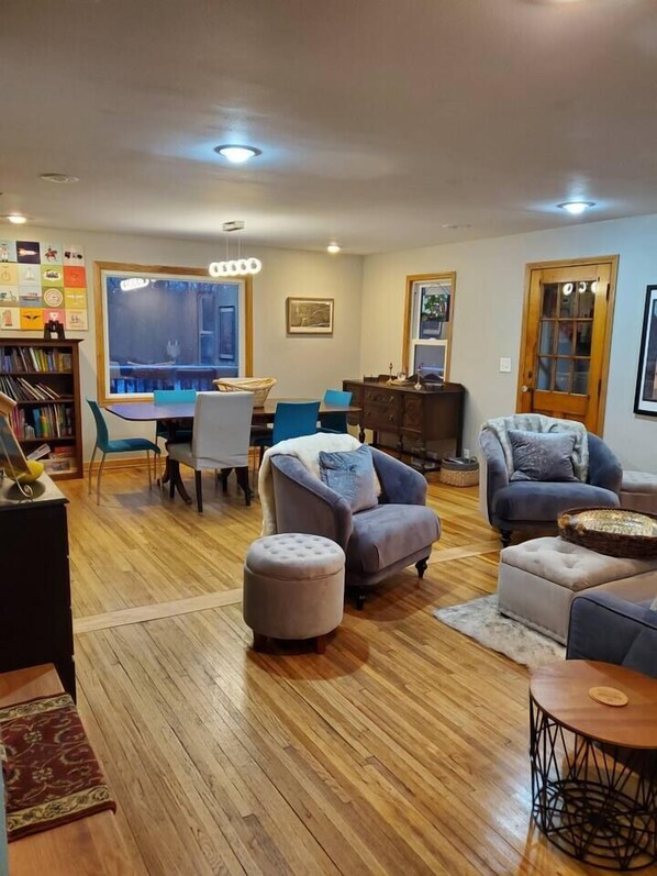 A spacious living room with a wooden floor, a dining area with a table and chairs, and a cozy seating area with armchairs and ottomans.