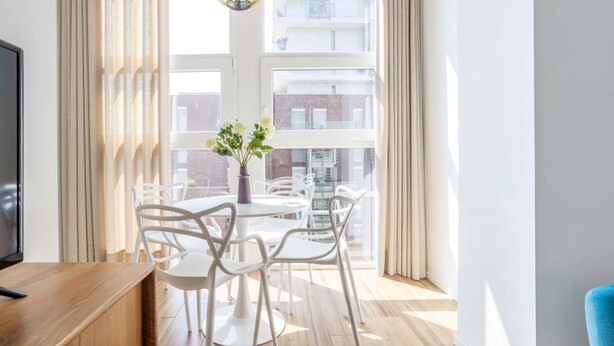 A modern room with a wooden desk, white chairs, a round table, a vase with flowers, and a large window with curtains. - Hostmate - Milano - Pirelli - Porta Nuova (Milano)
