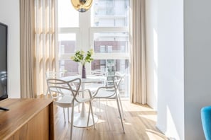 A modern room with a wooden desk, white chairs, a round table, a vase with flowers, and a large window with curtains.