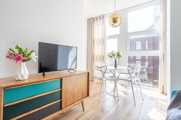 A modern living room with a TV on a wooden cabinet, a dining table with chairs, and a large window with curtains.
