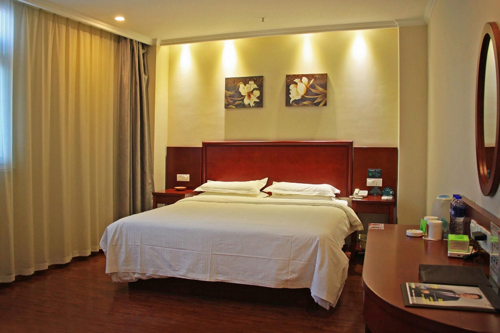 A hotel room with a large bed, wooden headboard, two wall-mounted paintings, a desk with a chair, and a nightstand with a lamp.