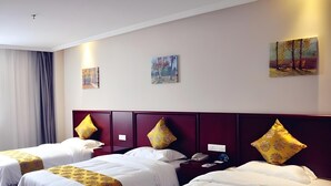 Habitación de hotel con tres camas, cada una con ropa blanca y almohadas decorativas. Hay cuadros en las paredes y una mesita de noche con teléfono y reloj.