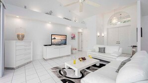 Un salon moderne avec un canapé blanc, une table basse et un téléviseur. On y trouve également un ventilateur de plafond, un meuble blanc et une porte avec un vitrage.