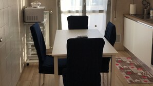 A small, well-lit kitchen with a dining table and chairs, a television mounted on the wall, and a large window with curtains.