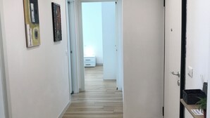 A hallway with wooden flooring, white walls, and two paintings. A door is visible on the right, and a bright room is seen in the distance.