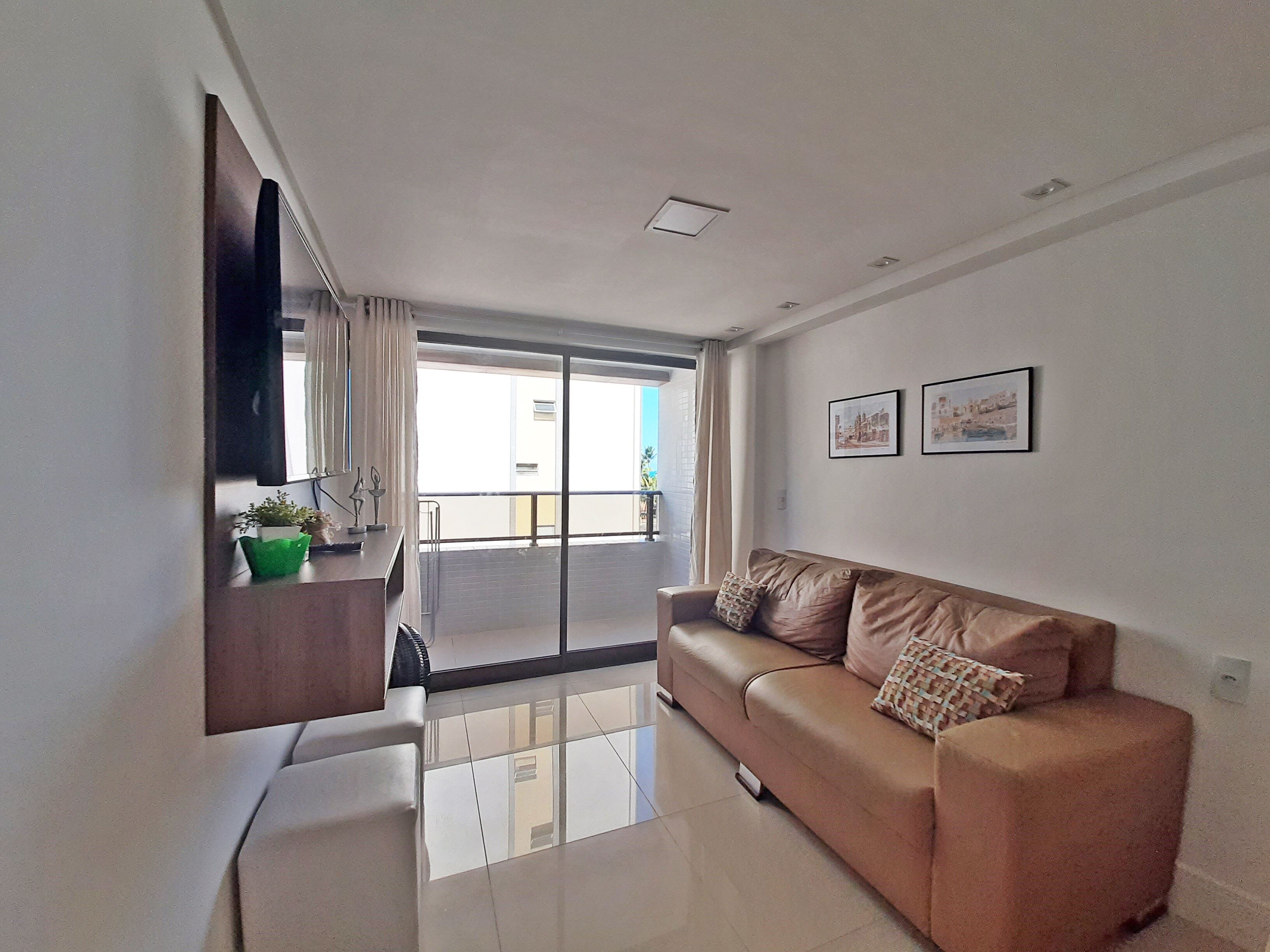A modern living room with a sofa, a flat-screen TV mounted on the wall, and a balcony with a view.