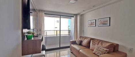 A modern living room with a sofa, a flat-screen TV mounted on the wall, and a balcony with a view.