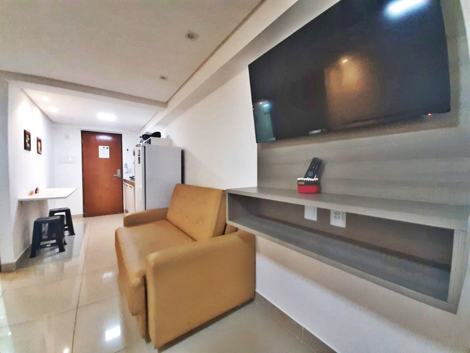 A living room with a sofa, a TV mounted on the wall, a refrigerator, and a door in the background.