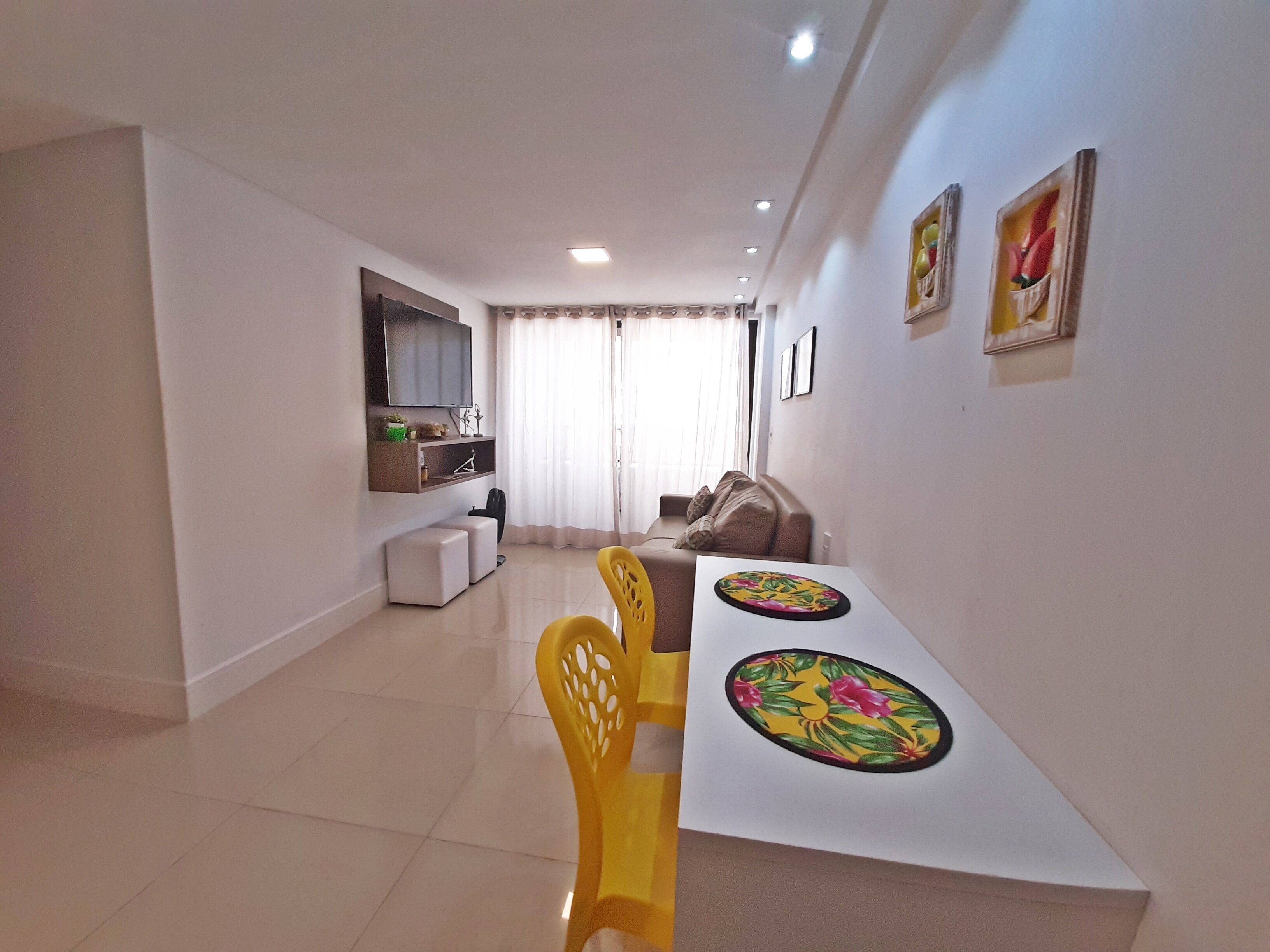 Una sala moderna con una mesa de comedor, sillas amarillas, un sofá, un televisor montado en la pared y una ventana con cortinas.