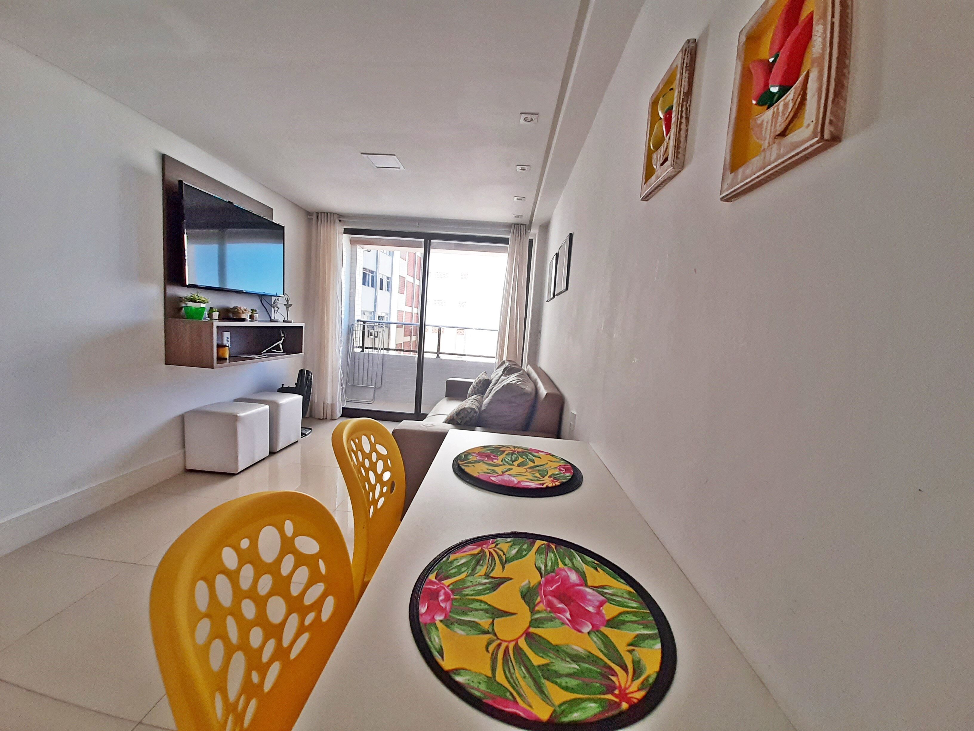 Ein modernes Wohnzimmer mit einer Couch, einem Esstisch mit Stühlen, einem an der Wand montierten Fernseher und einem Balkon mit Blick auf die Stadt.