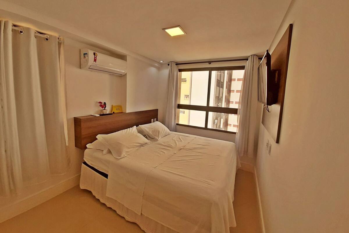 Habitación de hotel con cama, ventana con cortinas, aire acondicionado de pared y una pequeña repisa con un florero y un cartel.