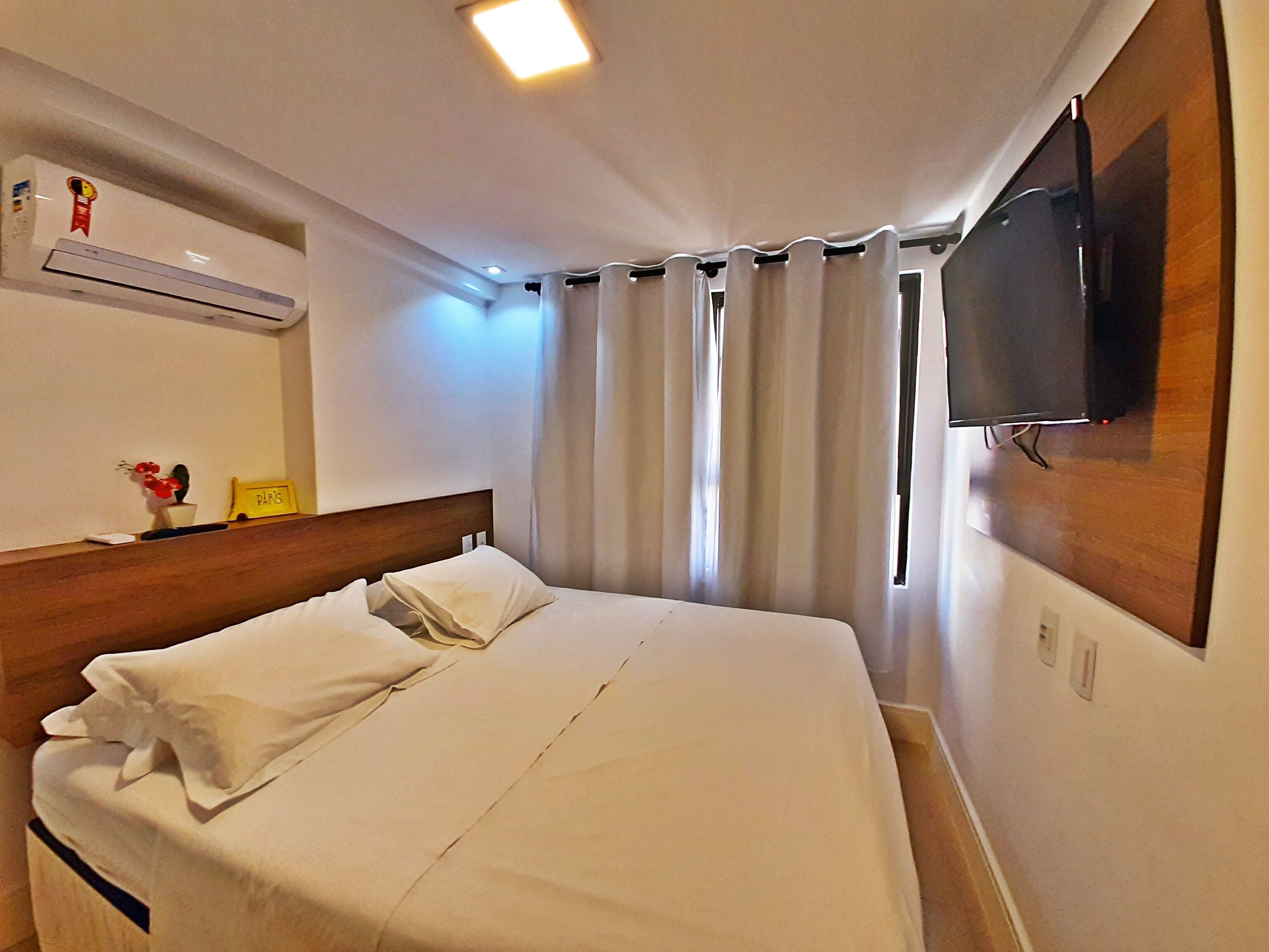 A hotel room with a bed, a mounted TV, an air conditioner, a small table with a plant, and a sign with the word "PARIS".