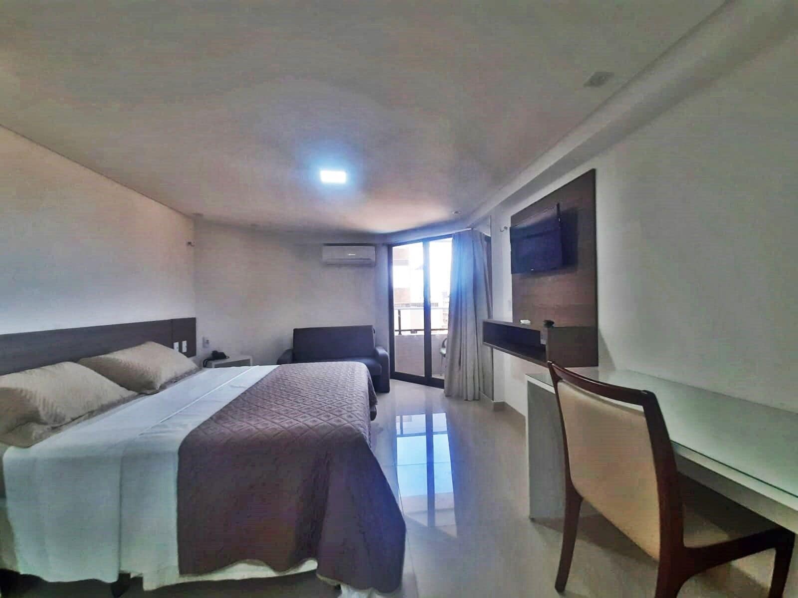 A hotel room with a bed, a desk, a chair, a TV, and an air conditioning unit.